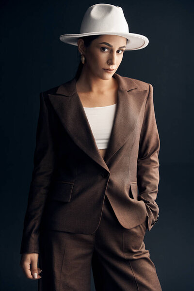 portrait photo on a dark background of a beautiful stylish girl in a brown suit, white top and white hat, she mysteriously looks at the camera with his brown eyes