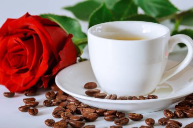 Beyaz kahve fincanı, kırmızı gül ve kahve çekirdekleri beyaz masaya saçılmış..