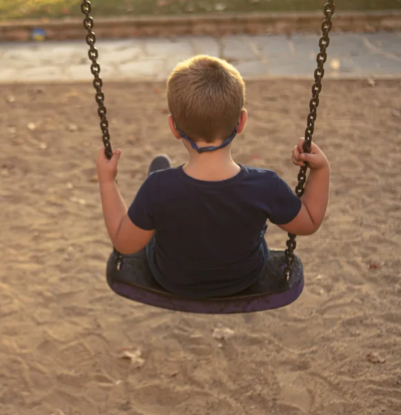 Blond boy with glasses swinging at sunset on a park swing — Stock Photo, Image