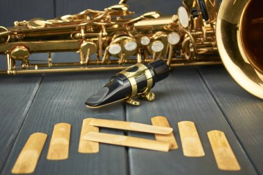 Metal kıskacı ve altın ayar donanımı ile saksafon sözlüğü, yedek sazlıklar ve gri ahşap arka planda enstrüman gövdesi
