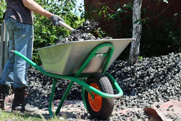 Человек с помощью лопаты загружает щебень в тачку. Закладка фундамента, проект строительства