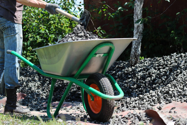 Человек с помощью лопаты загружает щебень в тачку. Закладка фундамента, проект строительства