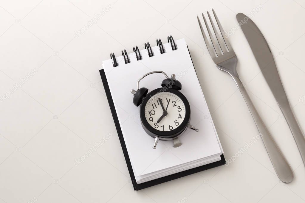 Clock on notepad with fork and knife for intermittent fasting and weight loss plan concept on white background