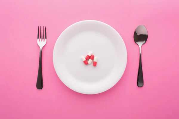 Étrend-kiegészítő tányéron villával és kanállal rózsaszín alapon — Stock Fotó