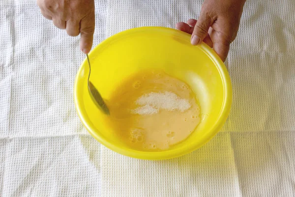 Female hands stir sugar with a raw egg in a yellow bowl, on a white towel background. Step by step cooking of pancakes. Step 3. Close-up hands of an elderly woman in the kitchen