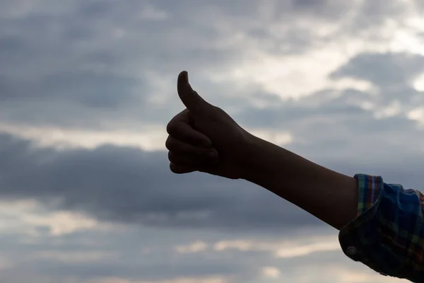 Silhouette of a childs hand makes a sign WELL on a background of cloudy sky. Outline of a hand with a raised thumb up. Good gesture, positive sign concept. Yes. Copy space