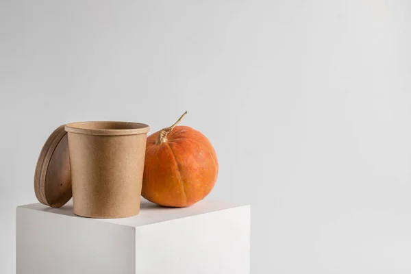 Empty paper cup on a white podium with a pumpkin on a white background. A brown container for soup, noodles, or other foods. An ecological product capable of degradation in a natural way.