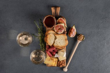 Salamlı peynirler, incir, ceviz, bal, tost ve tahta bir tahtanın üzerinde beyaz şarap. Karanlık bir taşta romantik bir randevu için yemek. Kopyalama alanı olan üst görünüm, koyu arkaplan.
