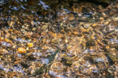 Işıl ışıl suda kayalar, berrak bir su deresinin suyunda yansıyan güneşli yansımalarla doğal arka planda zen meditasyonu, küçük dalgalar ve sağlıklı bir dağdaki ipeksi dalgalar görülüyor.