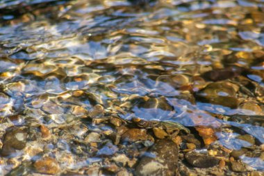 Işıl ışıl suda kayalar, berrak bir su deresinin suyunda yansıyan güneşli yansımalarla doğal arka planda zen meditasyonu, küçük dalgalar ve sağlıklı bir dağdaki ipeksi dalgalar görülüyor.