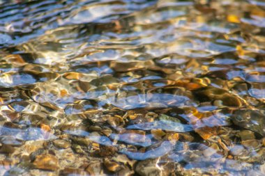 Işıl ışıl suda kayalar, berrak bir su deresinin suyunda yansıyan güneşli yansımalarla doğal arka planda zen meditasyonu, küçük dalgalar ve sağlıklı bir dağdaki ipeksi dalgalar görülüyor.