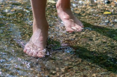 Küçük bir derede çıplak ayakla temiz suyla oynayan genç bir çocuk ayaklarını ve su kaynağını kullanarak ayak parmaklarını ve bacaklarını serinletiyor ve zen meditasyon atmosferindeki saf yaşam iksiriyle ferahlatıyor.