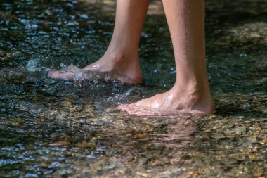 Küçük bir derede çıplak ayakla temiz suyla oynayan genç bir çocuk ayaklarını ve su kaynağını kullanarak ayak parmaklarını ve bacaklarını serinletiyor ve zen meditasyon atmosferindeki saf yaşam iksiriyle ferahlatıyor.