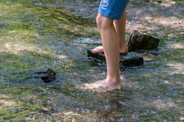 Küçük bir derede çıplak ayakla temiz suyla oynayan genç bir çocuk ayaklarını ve su kaynağını kullanarak ayak parmaklarını ve bacaklarını serinletiyor ve zen meditasyon atmosferindeki saf yaşam iksiriyle ferahlatıyor.
