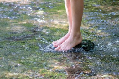 Küçük bir derede çıplak ayakla temiz suyla oynayan genç bir çocuk ayaklarını ve su kaynağını kullanarak ayak parmaklarını ve bacaklarını serinletiyor ve zen meditasyon atmosferindeki saf yaşam iksiriyle ferahlatıyor.