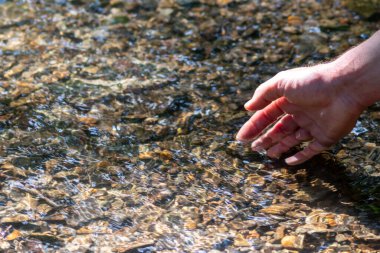 Avrupalı bir erkek el, ferahlatıcı bir zen atmosferde kristal gibi berrak bir suyun içinde ipeksi dalgalar ve yüzen dalgalar ve suda taş arayan güneşli yansımalarla.