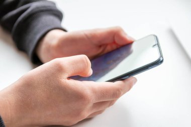 Avrupalı çocuk elleri siyah akıllı telefonu parmakları ve siyah ekranı beyaz masanın üzerinde tutuyor. Okuldaki araştırma ve mobil oyun için beyaz defterin önünde. Ya da çocukların ve yetişkinlerin oyun bağımlılığı için.