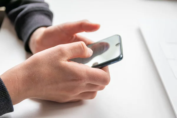 European child hands holding black smartphone with fingers and black screen on white desk infront of white notebook at school for research and mobile gaming or game addiction of kids and grown ups