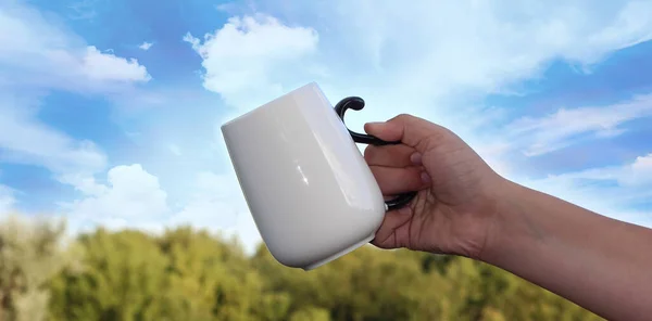 A white mug in a man\'s hand against a sky with clouds. Good morning.