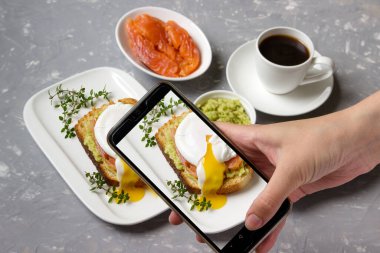 Somon füme ve avokado ile haşlanmış yumurta kahvaltısının fotoğrafı. Telefon kızın elindeydi..
