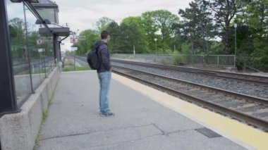 Tren istasyonunda eğitim bekleyen bir gencin 4K 'lık yüksek kaliteli videosu. Kanada 'daki GO tren istasyonu. Genç yakışıklı bir adam istasyonda duruyor ve elinde cep telefonu var. Mavi maske takıyor..