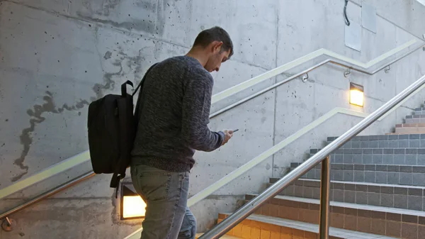 年轻人爬上车站的楼梯 手里拿着手机 背上背着背包 一个人在火车 公共汽车 地铁的飞机上赶时间 在车站上赶车 免版税图库照片