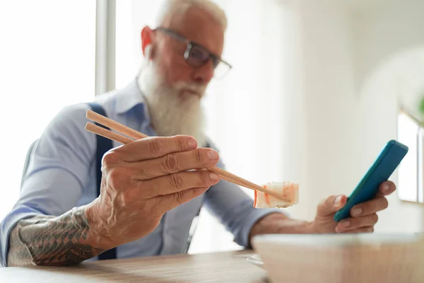 Portrait of modern old man looking on the phone in the office of house. Trendy architect eating sushi at lunch time. work, modern hipster concept. Focus on sushi - Image