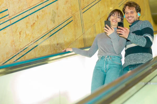 Capture happy moments. Joyful young loving couple looking a video. Happy Friends while going down the escalator and listening to music. New technology trends and friendship concept - Image.