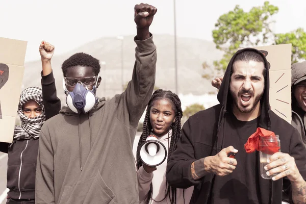 ガスマスクを持つ若い黒人男性は 公開デモで拳を上げて人々の群衆を行進します 黒人は正義と平等な権利のために路上で問題のデモを生きている — ストック写真