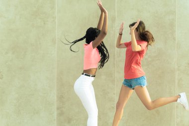 İzole edilmiş bir duvarın önüne atlayan neşeli genç arkadaşların görüntüsü. İki mutlu genç kadın el ele tutuşuyor. Resim.