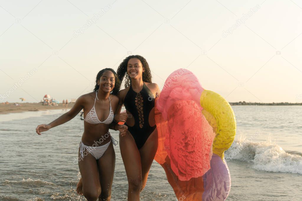 Happy crazy friends running with ice cream inflatable mat. Young girls having fun near of ocean. Summer, adventure, travel lifestyle and vacation concept. Image