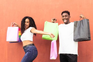 Alışveriş torbaları olan mutlu genç bir çift, arka plan duvarı kırmızıda izole edilmiş. Alışveriş merkezindeki alımdan sonra arkadaşlar çok mutlu oldular. Tatil, arkadaşlık ve satın alma kavramı.