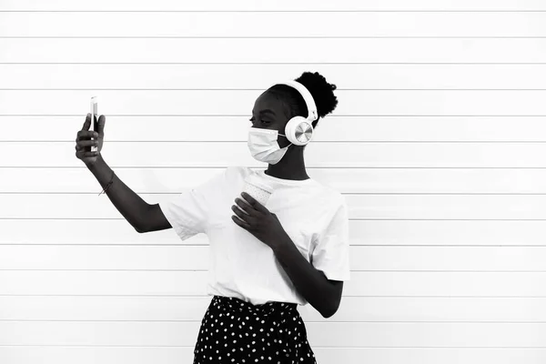 若いアフリカ人女性が携帯電話を使って自撮りや音楽を聴いている コロナウイルスの予防のためにフェイスマスクをしている黒の女の子 技術とウイルスの概念 白黒写真 — ストック写真