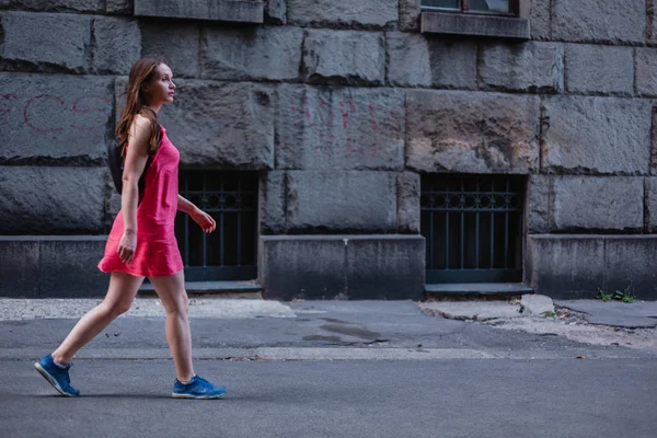 Side view of woman in pink dress with backpack walking by street