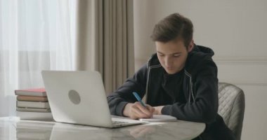 Genç çocuk evde ders veriyor. Ödevi öğrenci yazıyor. Çocuk öğreniyor. Çocuk dizüstü bilgisayar kullanıyor. Öğrenci not defterine kalemle yazar..