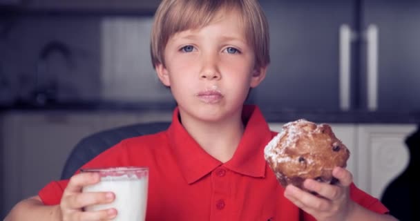 小男孩喝牛奶和蛋糕 7岁的男孩带着面包和牛奶吃早餐 可爱的快乐的孩子早上吃健康的食物 金发男孩和一个美味的甜松饼 实时录像 — 图库视频影像