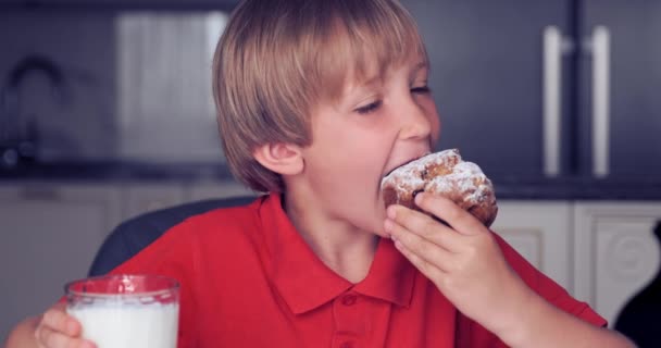 小男孩喝牛奶和蛋糕 7岁的男孩带着面包和牛奶吃早餐 可爱的快乐的孩子早上吃健康的食物 金发男孩和一个美味的甜松饼 慢动作 — 图库视频影像