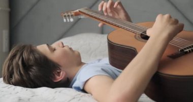Evde akustik gitar çalan genç bir adam. Tahta gitarlı genç bir çocuk kanepede yatıyor. Gitar çalan çocuk. Genç adam klasik gitarla çalıyor. Gerçek zamanlı görüntüler. 