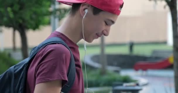 在街上走来走去的青少年 用耳机听音乐 戴着红色棒球帽的学生在户外用耳机听音乐 戴耳机的少年外出 城市场景 — 图库视频影像