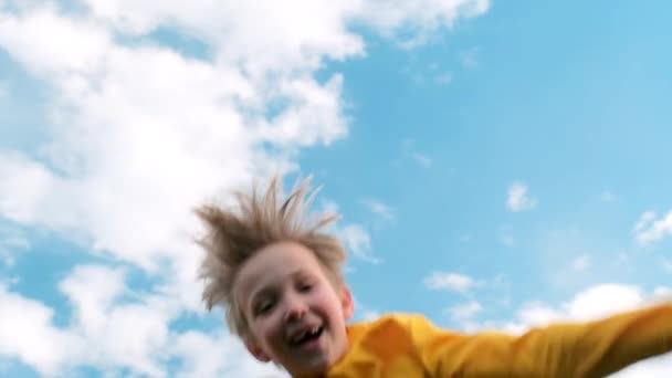幸せな子供は青い空に対してトランポリンで非常に高いジャンプします トランポリンで金髪の男の子がジャンプします トランポリンでジャンプする陽気な男の子のスローモーションの下のビュー 子供の幸せ — ストック動画