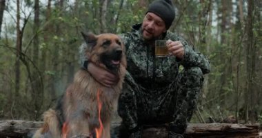 Yetişkin Adam, köpeğiyle birlikte ateşte dinleniyor. Yalnız gezgin orman yangınında Alman çoban köpeği ile dinleniyor ve çay içiyor. İnsan, ormandaki yangında bir köpeği okşar.. 