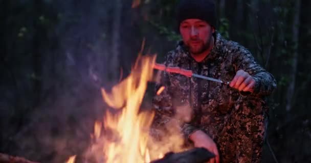 男人在森林里的火堆上煎香肠 旅行者在森林里生火做饭 夏天的一个晚上 一个成年人坐在篝火边做饭 人类在森林中休息 — 图库视频影像