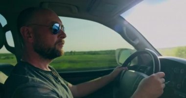 Adam gün batımında arabayı sürüyor. Arabayı kullanan yetişkin sakallı adam. Genç yetişkin bir çocuk araba kullanıyor. Gerçek zamanlı. Arabanın ön camında güneş ışınları parlıyor. Araba süren adama yan bakış.. 