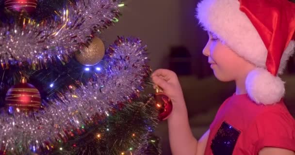 明るいおもちゃやガーランドとクリスマスツリーで笑顔の男の子の4Kスローモーションビデオ サンタの帽子の男の子はクリスマスツリーにクリスマスのおもちゃを掛けます 幸せな子供は新年の準備をしています — ストック動画