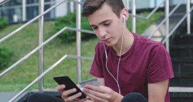 Gençler internet üzerinden kredi kartı ve cep telefonu ile alışveriş yapıyorlar. Gülümseyen genç adam açık havada, banka kartlı cep telefonuyla online alışveriş yapıyor. Çocuk akıllı telefon ve kredi kartı kullanıyor. Gerçek zamanlı..