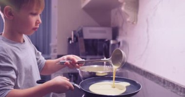 Mutfakta yemek pişiren 8 yaşındaki bir çocuk. Çocuk kepçeden tavaya krep kızartmak için karışımı dolduruyor. Çocuk krep karışımını çırpıcıyla çırpıyor. Ağır çekim. Çocukluk kavramı