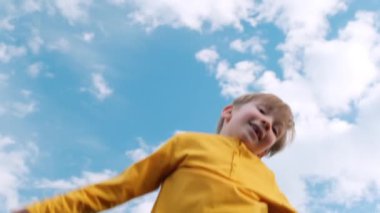 Mutlu çocuk mavi gökyüzüne karşı trambolinde çok yükseğe zıplar. Sarışın çocuk trambolinde zıplıyor. Trambolinde zıplayan neşeli bir çocuğun yavaş çekim alt görüntüsü. Çocukların mutluluğu. 