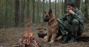 Yalnız gezgin, orman yangınında Alman çobanıyla sıcak bir içecekle dinleniyor. Köpekli bir adam ormanda ateşin başında oturur. Yetişkin Adam köpeğiyle birlikte ateşte dinleniyor.