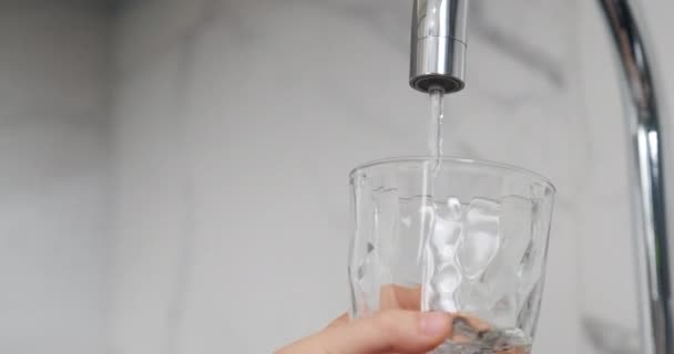 孩子的手拿着一个透明的杯子杯 盛满了清洁的水 把装满水的杯子关上 小孩的手在厨房水龙头边拿着透明的杯子 水龙头里有水流 — 图库视频影像