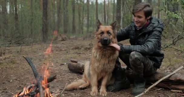 小男孩带着大狗坐在森林的火边 快乐的家伙在户外摸他的狗 一个快乐的高加索年轻人和他的德国牧羊人在大自然的火边 — 图库视频影像